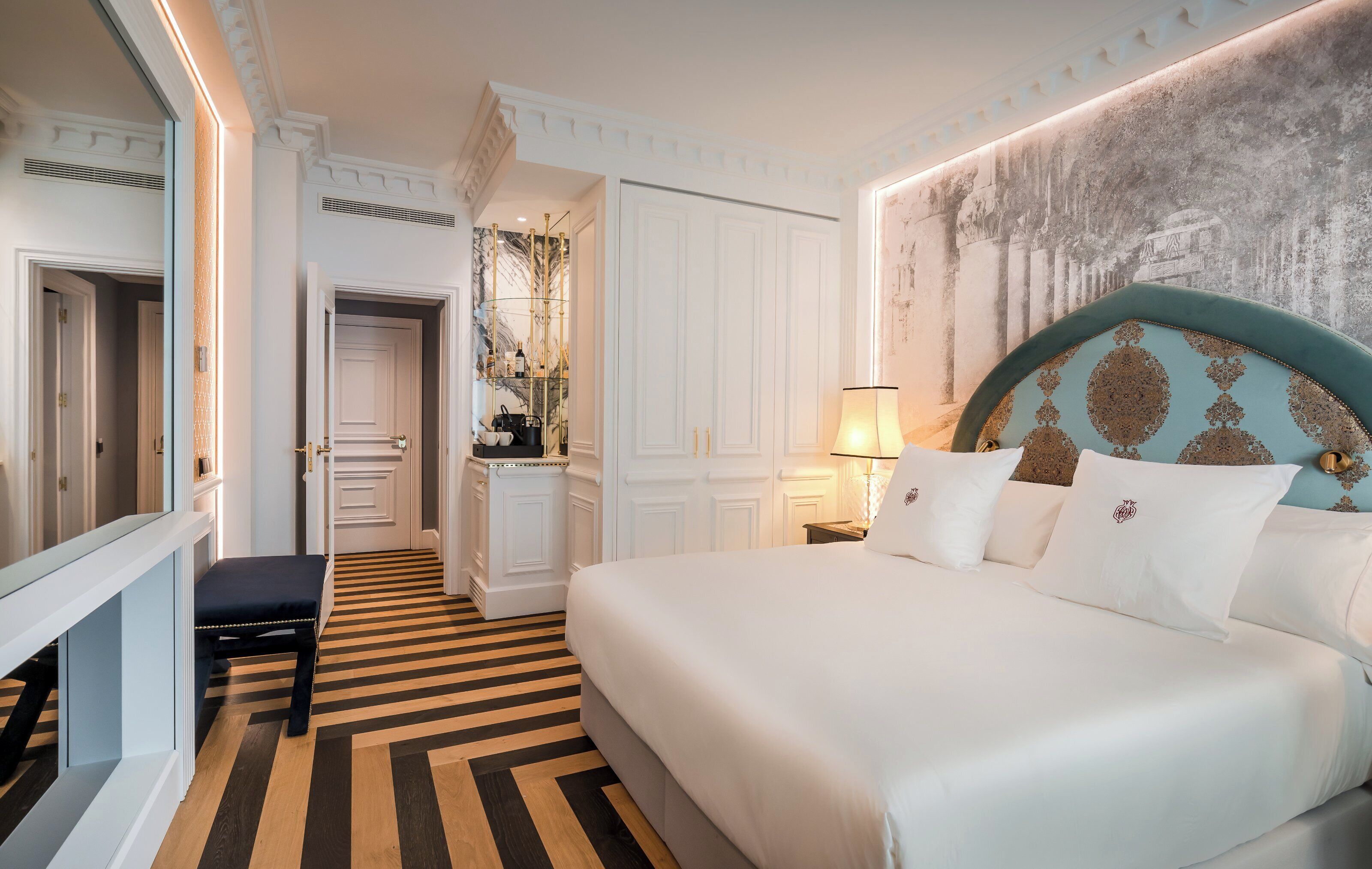 A hotel room with a large bed, a bench, a desk, and a mirror.