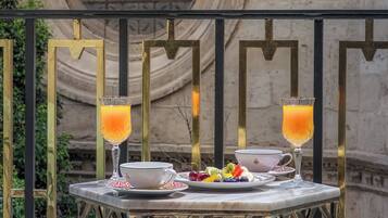 A table with a white cup, saucer, and a plate of fruit, two glasses of orange juice, and a candle on a balcony.