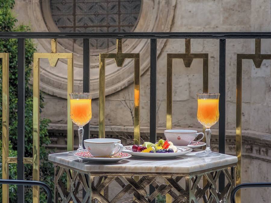 A table with a white cup, saucer, and a plate of fruit, two glasses of orange juice, and a candle on a balcony.