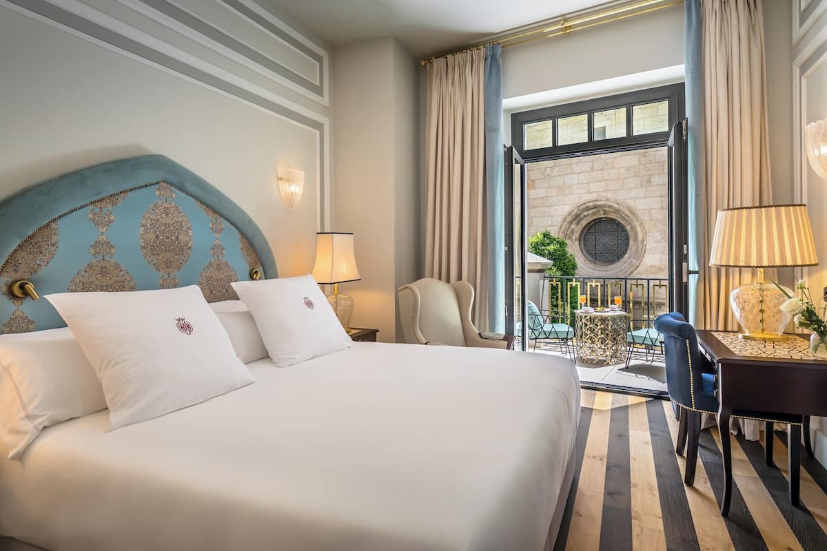A hotel room with a large bed, a desk, a chair, and a view of a courtyard through an open door.