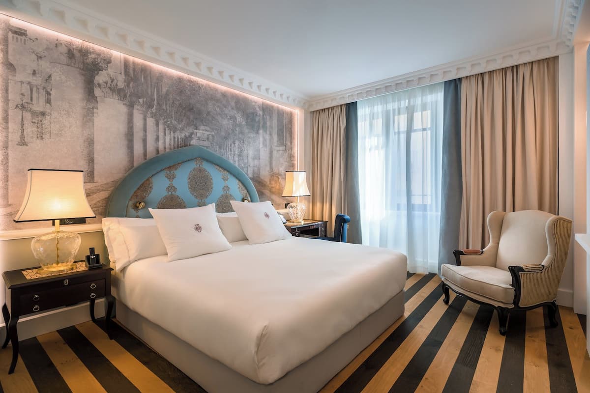 A hotel room with a large bed, two bedside tables with lamps, a chair, a large window with curtains, and a textured wall.