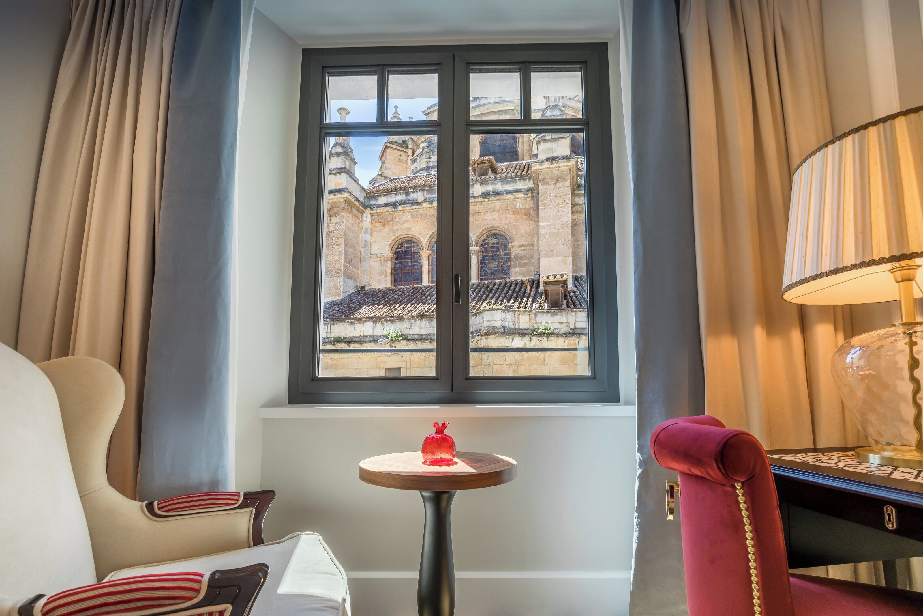 Habitación de hotel con un ventanal grande, una mesita redonda con un objeto rojo, una lámpara y una silla con cojines a rayas.
