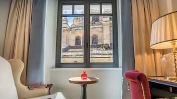 Habitación de hotel con un ventanal grande, una mesita redonda con un objeto rojo, una lámpara y una silla con cojines a rayas.