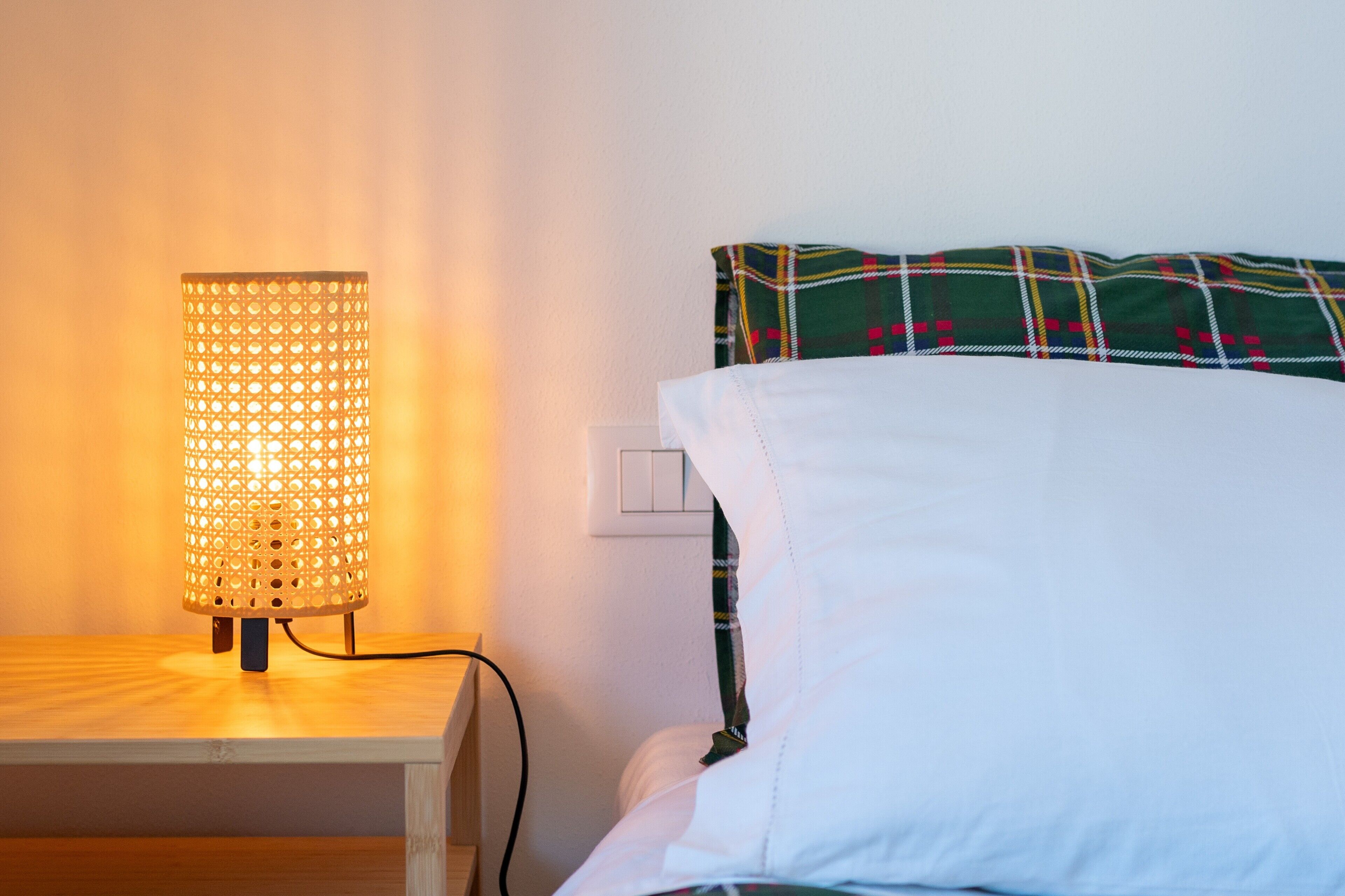 Una cama con un cojín escocés y una lámpara de noche en una mesita de madera.