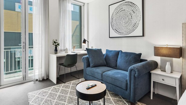 Un salon moderne avec un canapé bleu, une petite table basse ronde, un bureau avec une chaise et une grande fenêtre avec des rideaux.