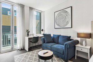 A modern living room with a blue sofa, a small round coffee table, a desk with a chair, and a large window with curtains. - Cozy 1-bed Studio in the Heart of Melbourne (Melbourne)