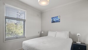A bedroom with a large bed, a nightstand with a lamp, a window with blinds, and a painting on the wall. - Central 2-bed Pad With Tennis Court, (Melbourne)