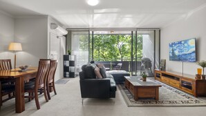 A modern living room with a sofa, coffee table, and dining area. There is a large window with blinds, a painting on the wall, and a ceiling light. - Central 2-bed Pad With Tennis Court, (Melbourne)