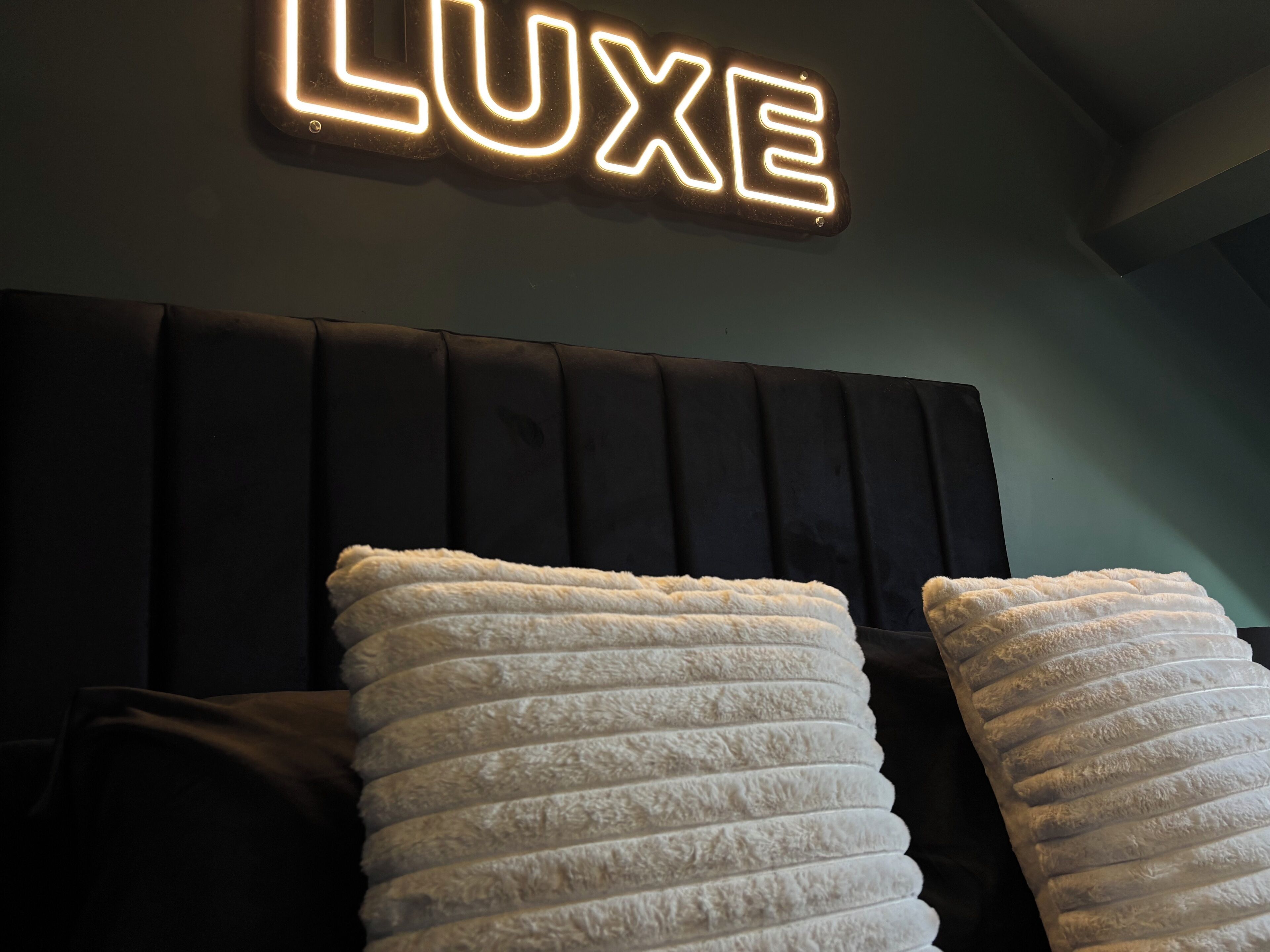 Uma cama com cabeceira preta e duas almofadas brancas. Acima da cama, um letreiro de neon com a palavra 'LUXE'.