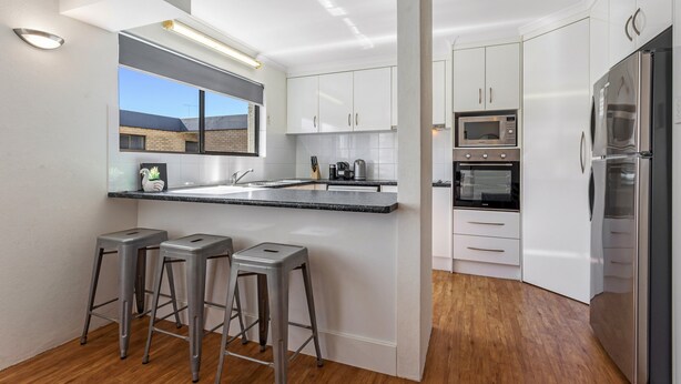 A modern kitchen with white cabinets, stainless steel appliances, a central island with stools, and a view of the outside. - Cosy 2-bedroom Seaside Unit Steps From Beach (Sunshine Coast)