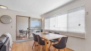 A modern dining area with a wooden table and black chairs, a leather sofa, and large windows with blinds. - Cosy 2-bedroom Seaside Unit Steps From Beach (Sunshine Coast)