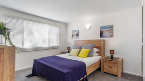 A bedroom with a bed, bedside tables, a nightstand, a window with blinds, and two wall-mounted picture frames. - Cosy 2-bedroom Seaside Unit Steps From Beach (Sunshine Coast)