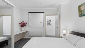 Una habitación de hotel moderna con una cama grande, una mesita de noche con lámpara, un escritorio pequeño, una silla y un espejo amplio.