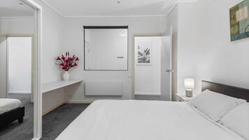 A modern hotel room with a large bed, a bedside table with a lamp, a small desk, a chair, and a large mirror.