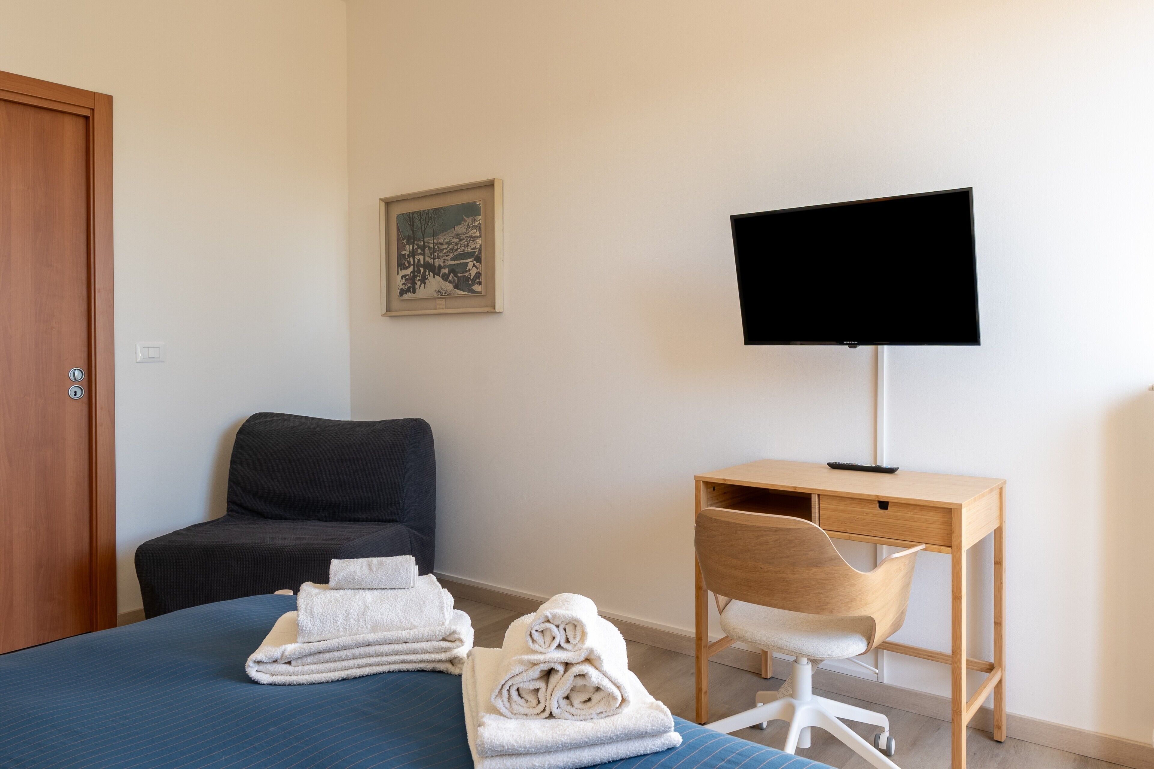 A hotel room with a bed, a desk, a chair, a TV, and a painting on the wall.