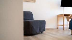 Ein Zimmer mit grauem Sofa, einem Holznachttisch und einem an der Wand befestigten Fernseher.