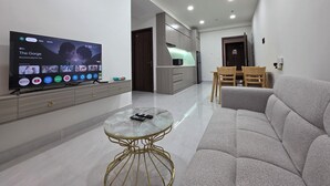 A modern living room with a flat-screen TV, a sofa, a glass coffee table, and a kitchen area in the background.