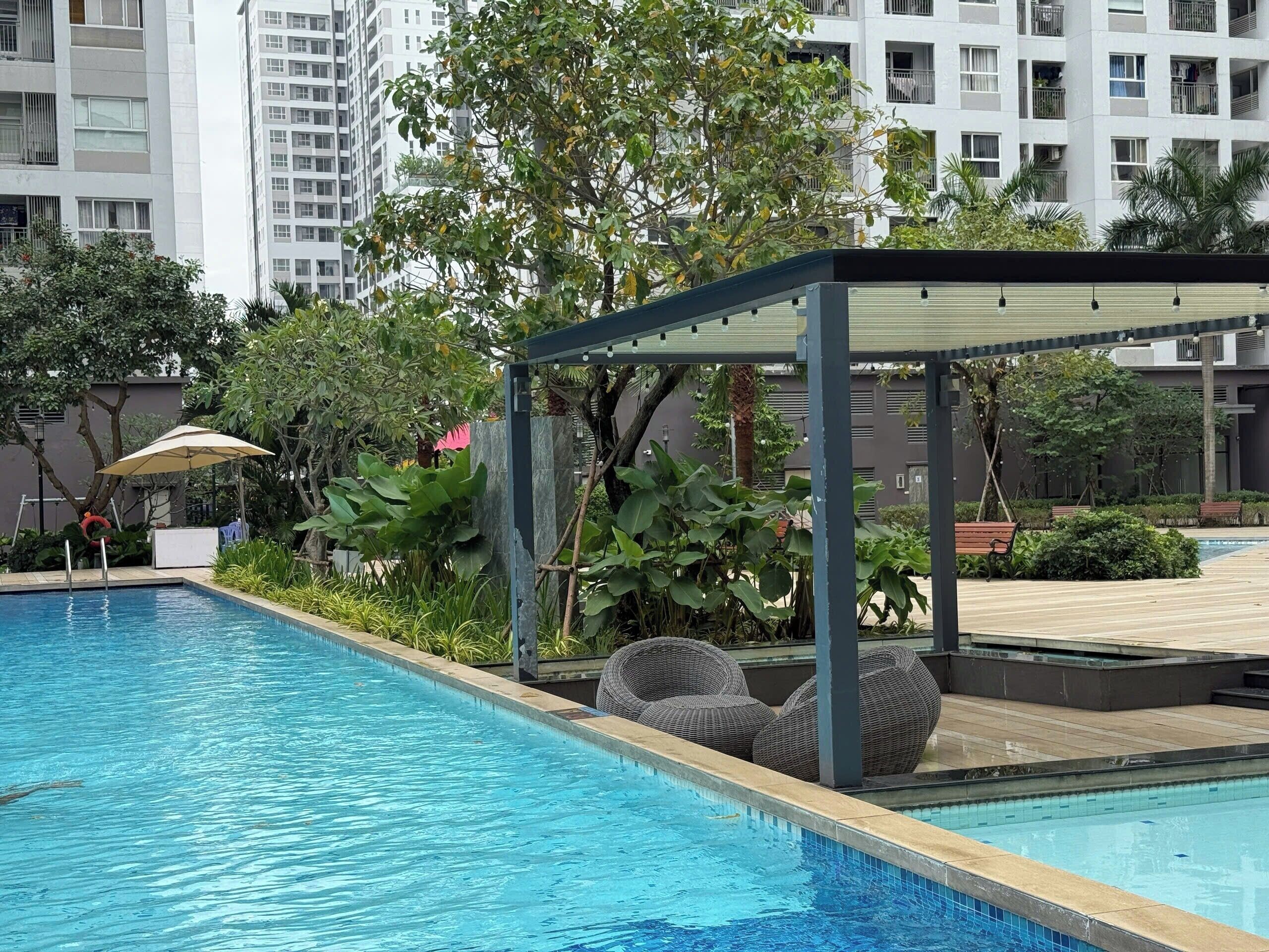 Piscina com área de estar coberta, rodeada por vegetação e edifícios modernos.