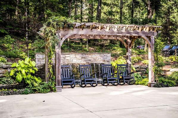 A wooden pergola with seating, surrounded by lush greenery and a stone wall.