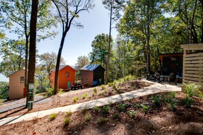 Un chalet rustique, accessible par un sentier de terre qui mène à une terrasse en bois, le tout entouré d’une forêt dense.