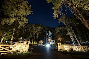 Una entrada protegida por un muro de piedra, con un letrero que indica vigilancia, y un edificio con ventanas iluminadas al fondo.