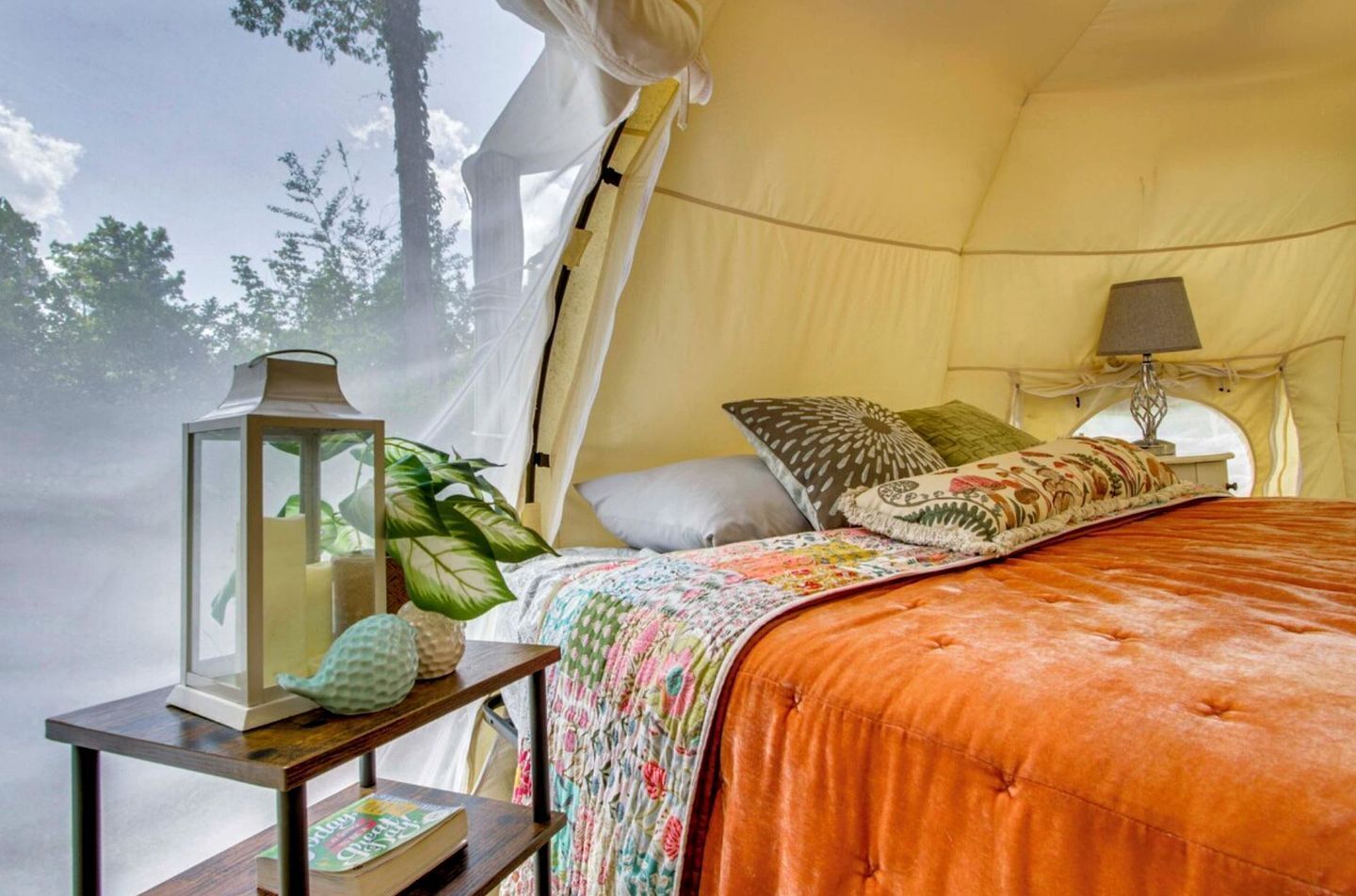 A tented room with a bed, a table with a lantern, and a window with a view of trees.