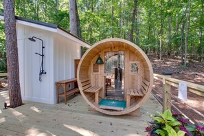 A wooden sauna structure with benches and a shower area, surrounded by a deck and forest.