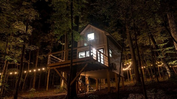 A treehouse with a balcony, surrounded by trees, and illuminated by string lights. - Amazing Tree House Perfect for a Secluded Getaway in Alabama (Scottsboro)