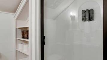 Une salle de bain avec une douche à parois de verre, des étagères blanches où sont posées des serviettes et une petite fenêtre.