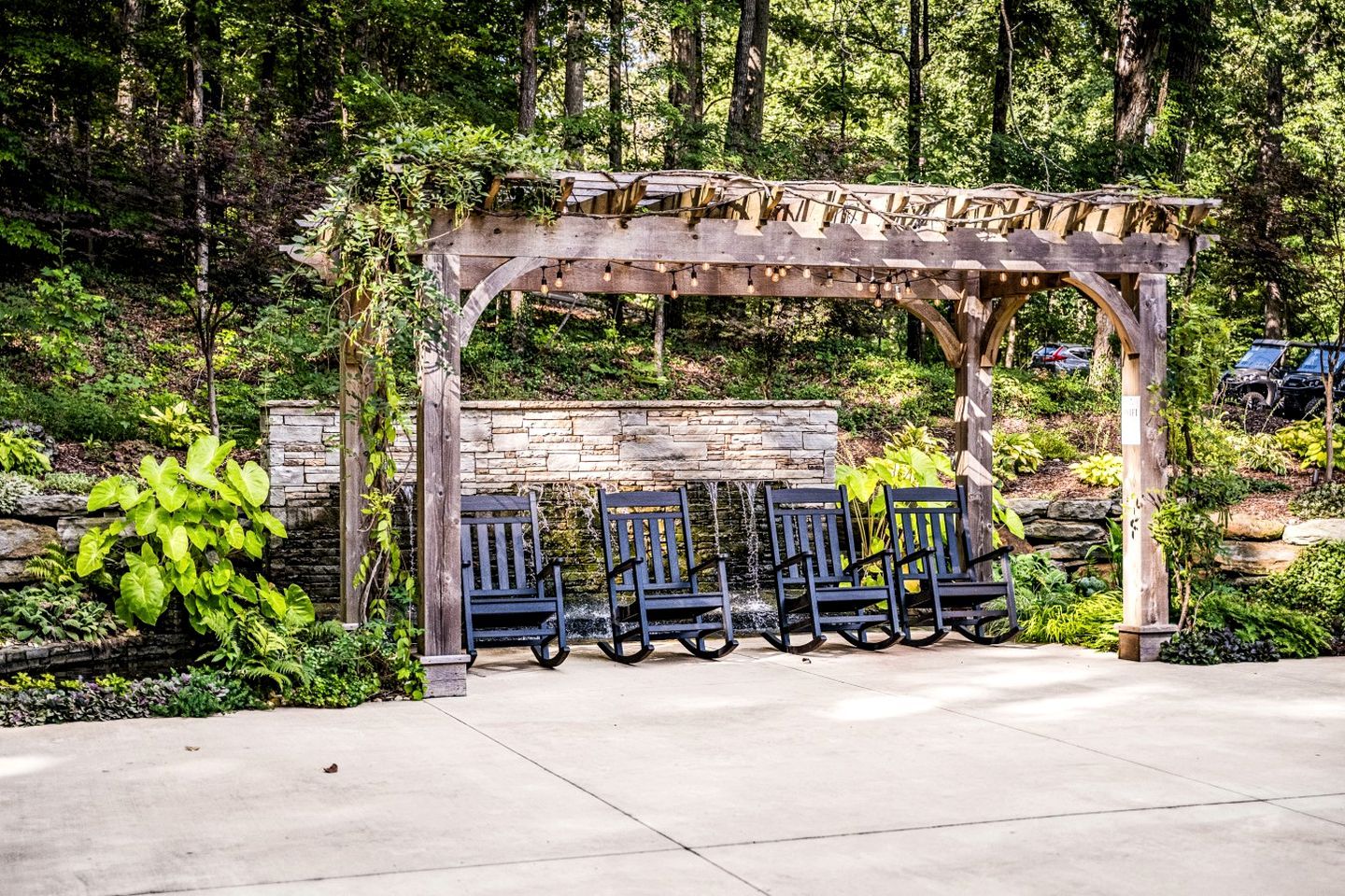 Une pergola en bois avec des sièges, entourée d’une végétation luxuriante et d’un mur de pierre.