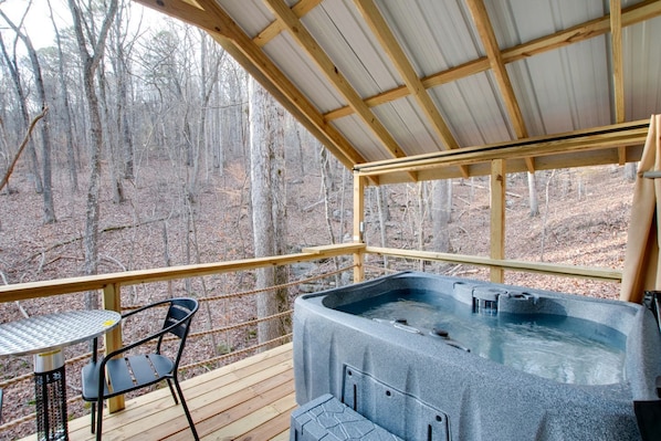 A hot tub on a wooden deck with a metal table and chair, surrounded by a metal railing, overlooking a forest.