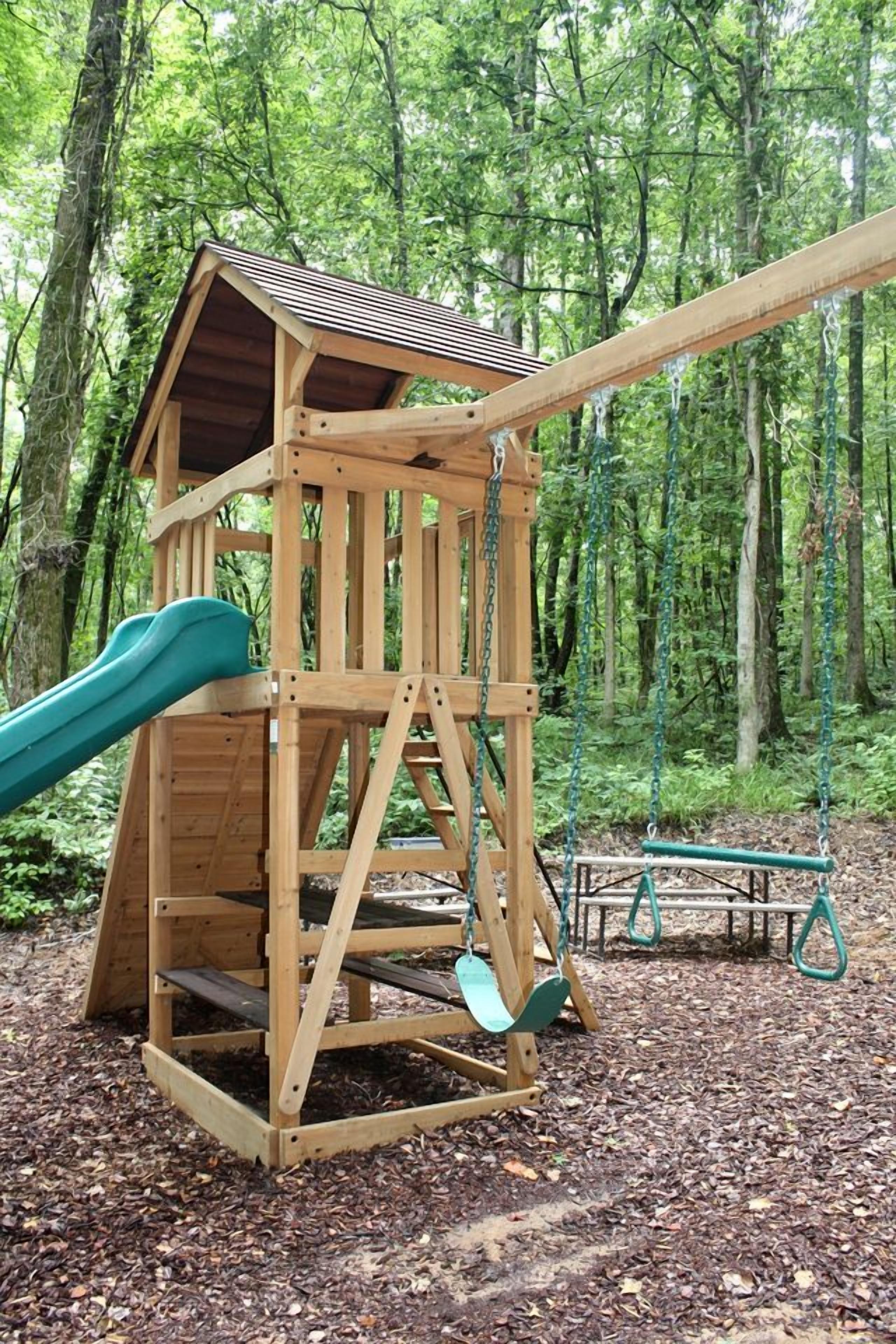 A wooden play structure with a slide and swings in a wooded area.