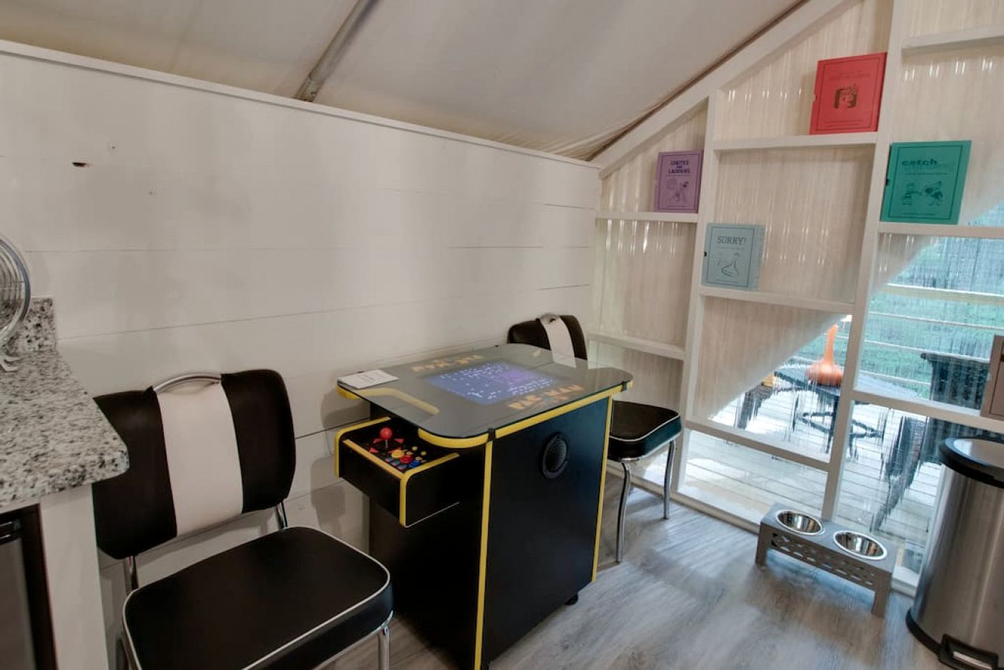 A room with a gaming table, two chairs, a pet feeding station, and a bookshelf with books.