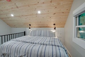 Un dormitorio con techo inclinado, una cama con ropa de cama a rayas, dos lámparas fijadas a la pared y una ventana con vistas.