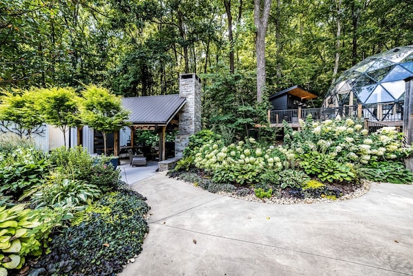 A modern house with a stone chimney, a covered patio, and a garden with various plants.