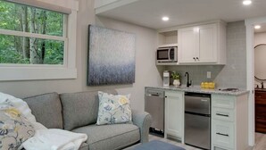 A modern kitchen with white cabinets, a microwave, and a dishwasher. There's a grey sofa with floral cushions and a bench with books. - Pet-Friendly Tree House Great for a Relaxing Escape in Alabama (Scottsboro)