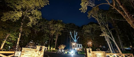 A gated entrance to a property with a stone wall, a sign indicating surveillance, and a building with illuminated windows in the background.