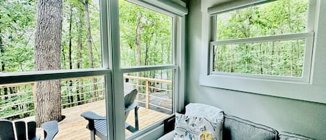 A room with a gray sofa, a patterned pillow, and a view of a wooden deck and trees through large windows.