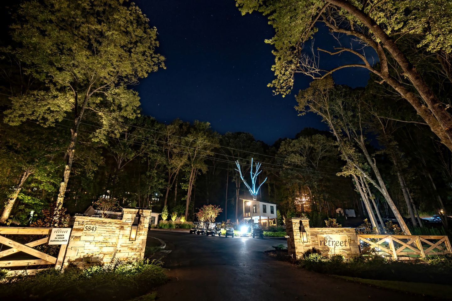 A gated entrance to a property with a stone wall, a sign indicating surveillance, and a building with illuminated windows in the background.