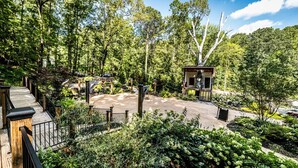 A wooden deck with a view of a forested area, a gazebo, and a paved area with a statue.