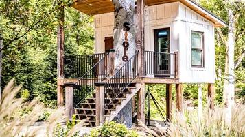 Une cabane dans les arbres avec une structure extérieure en bois, un balcon et un escalier menant à l’intérieur.