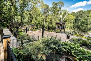 A wooden deck with a view of a forested area, a gazebo, and a paved area with a statue.