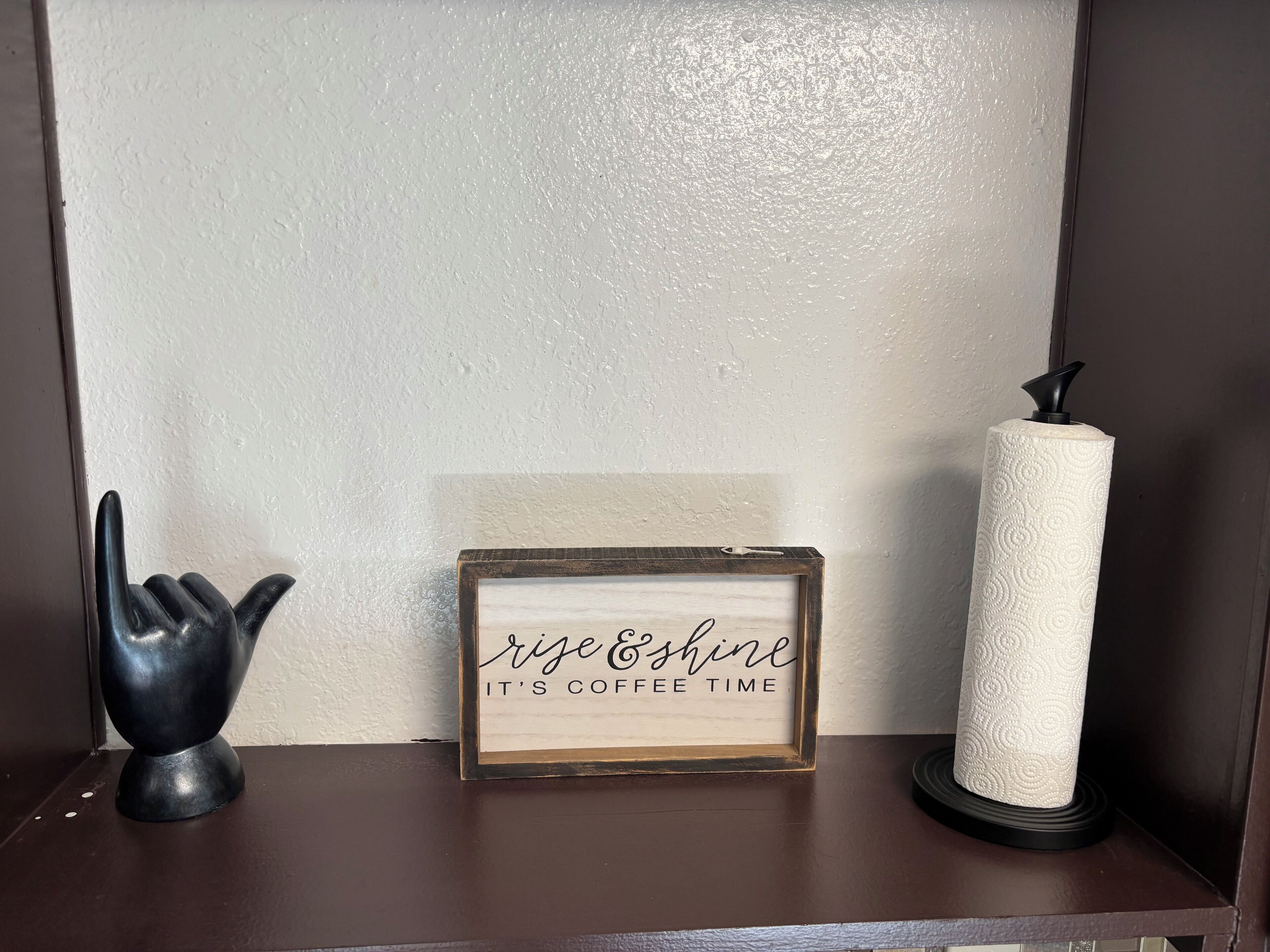 Ein gerahmtes Schild mit dem Aufdruck 'rise & shine it's coffee time' auf einem Regal.