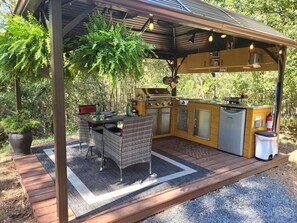 Zona cuina exterior amb forlla, rentapeus i armaris d'emmagatzematge, envoltada d'una terrassa de fusta i plantes en testos.