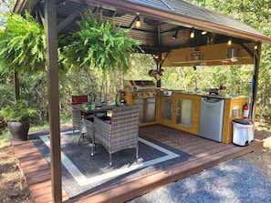 An outdoor kitchen area with a grill, sink, and storage cabinets, surrounded by a wooden deck and potted plants.