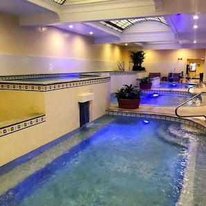 An indoor swimming pool with a skylight ceiling, tiled walls, and potted plants.
