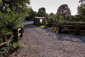 Un chemin de gravier mène à une construction rustique en bois dotée d’une porte en verre, le tout étant entouré de verdure et d’une clôture en bois.