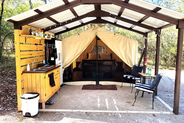 Un área de cocina al aire libre techada con pileta, armarios de almacenamiento y una zona para dormir cubierta con carpa, cama y asientos.