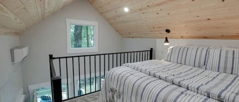 A bedroom with a sloped ceiling, a window, a bed with striped bedding, a black railing, and a small table with chairs outside.