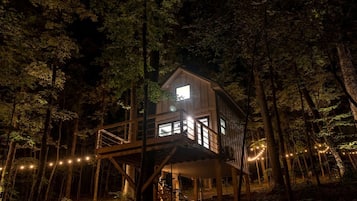 A treehouse with a balcony, surrounded by trees, and illuminated by string lights.